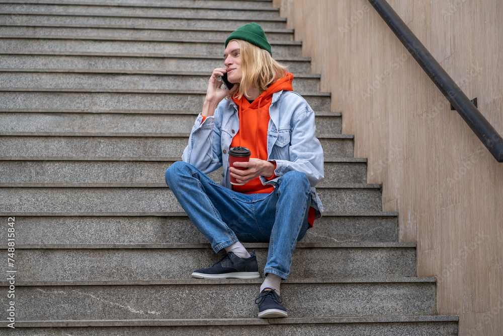 Joyful student guy smiling talking on cellphone sitting on city staircase with takeaway coffee. Contented young hipster man have nice chatting conversation on street with girlfriend on mobile phone. Joyful student guy smiling talking on cellphone sitting on city staircase with takeaway coffee. Contented young hipster man have nice chatting conversation on street with girlfriend on mobile phone.