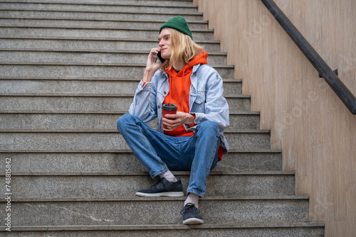 Joyful student guy smiling talking on cellphone sitting on city staircase with takeaway coffee. Contented young hipster man have nice chatting conversation on street with girlfriend on mobile phone. © DimaBerlin