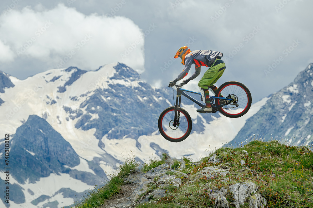 Mountain biker defies gravity, soaring mid-air against backdrop of snow-capped peaks and lush greenery. Ideal for adventure, sports, and nature themes