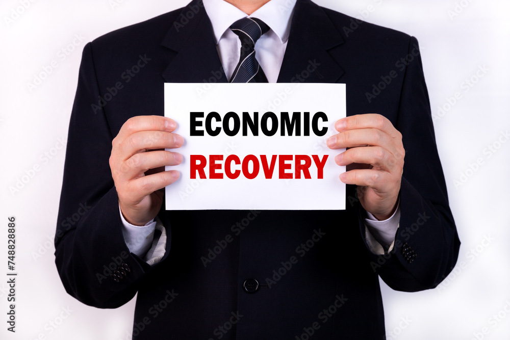 A businessman is holding a tablet with the text Economic Recovery in front of him in his hands. Business concept A businessman is holding a tablet with the text Economic Recovery in front of him in his hands. Business concept