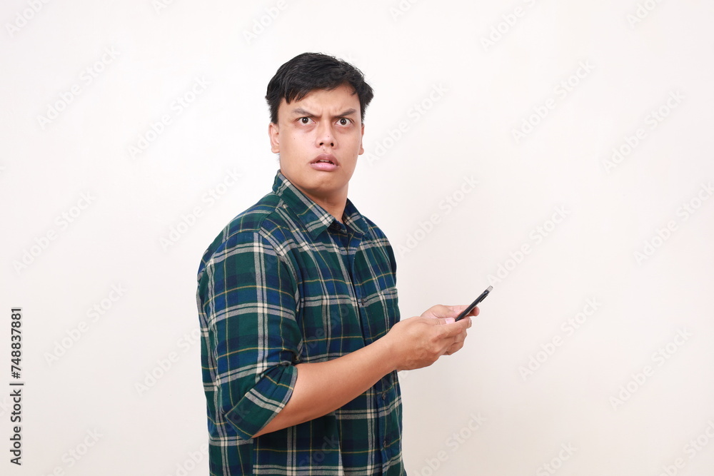 Young asian man holding a cell phone with angry face expression Young asian man holding a cell phone with angry face expression
