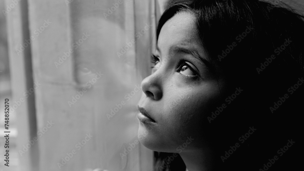 One unhappy somber little girl feeling depressed during hard times. Close-up child's face and hand leaning on glass window feeling lonely and solitude in black and white, monochromatic