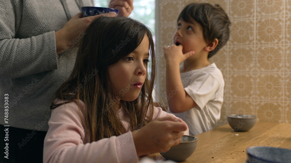 Pensive little girl eating yogurt from bowl in kitchen with mother and little brother in the background. Domestic authentic lifestyle scene of family eating breakfast early in the morning Pensive little girl eating yogurt from bowl in kitchen with mother and little brother in the background. Domestic authentic lifestyle scene of family eating breakfast early in the morning