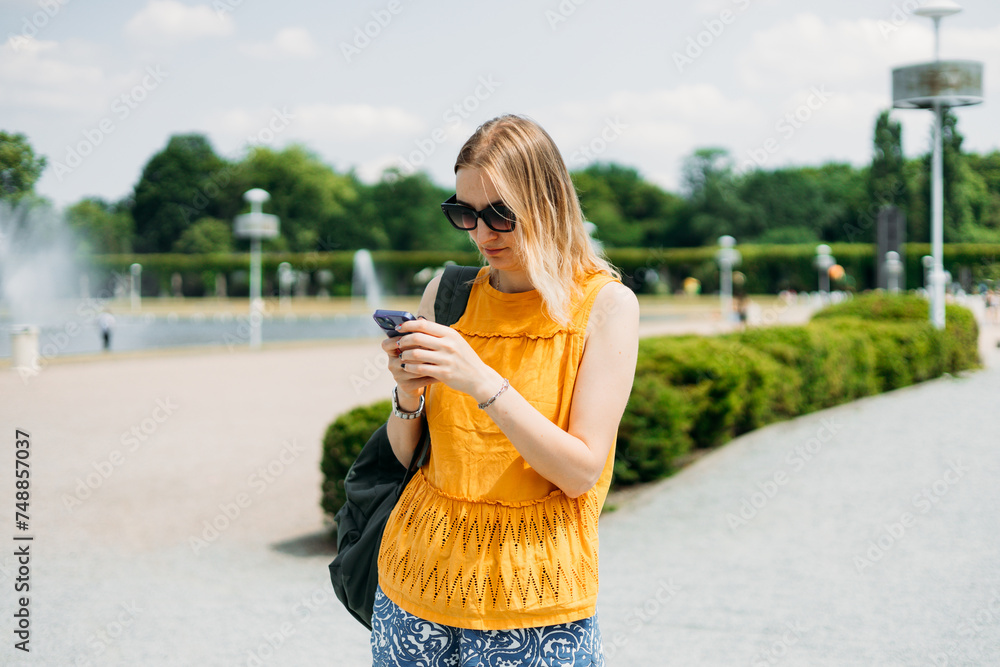 Happy cheerful young blond woman with backpack walking on city street checks her smartphone. Portrait of beautiful 30s girl using smartphone outdoors. Urban lifestyle concept. Summer time