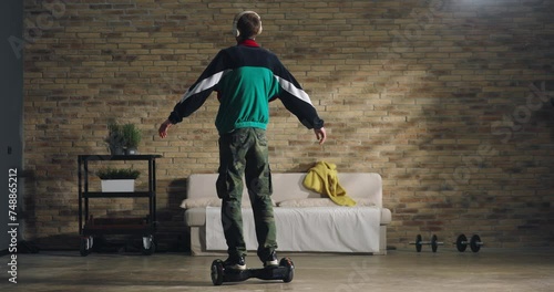 Young teenager wearing headphones is having fun to listen his favorite music playlist and feeling free with open arm while rides technological gadget hoverboard at home. photo