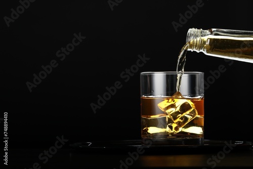 Pouring whiskey from bottle into glass with ice cubes at table against black background, space for text