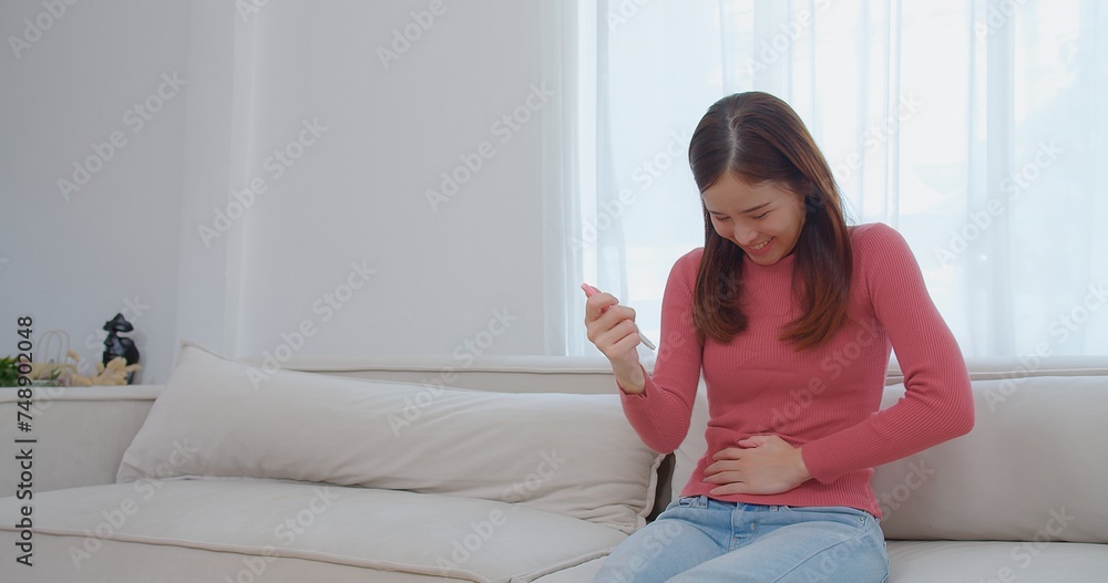 Ecstatic young asian woman in pink turtleneck holding looking at a pregnancy test and surprise exciting reaction laughing smile with happiness on a white couch, celebrating good news, a moment of joy Ecstatic young asian woman in pink turtleneck holding looking at a pregnancy test and surprise exciting reaction laughing smile with happiness on a white couch, celebrating good news, a moment of joy