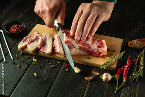 Cutting lard with a knife in the chef hand to prepare a delicious snack on the kitchen table with pepper and garlic. Fatty food concept for weight gain