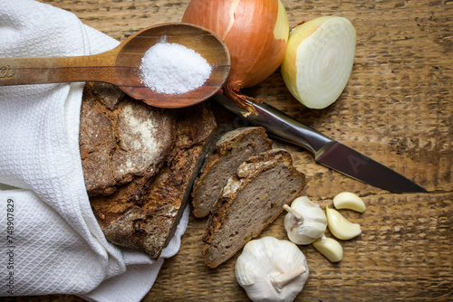 bread placed on a cloth with a knife, garlic, onion, salt and a wooden spoon on a wooden background