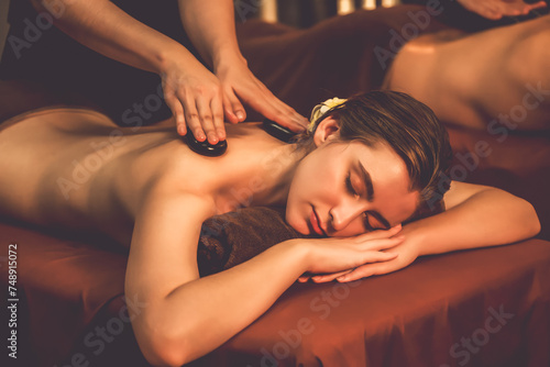 Hot stone massage at spa salon in luxury resort with warm candle light, blissful couple customer enjoying spa basalt stone massage glide over body with soothing warmth. Quiescent