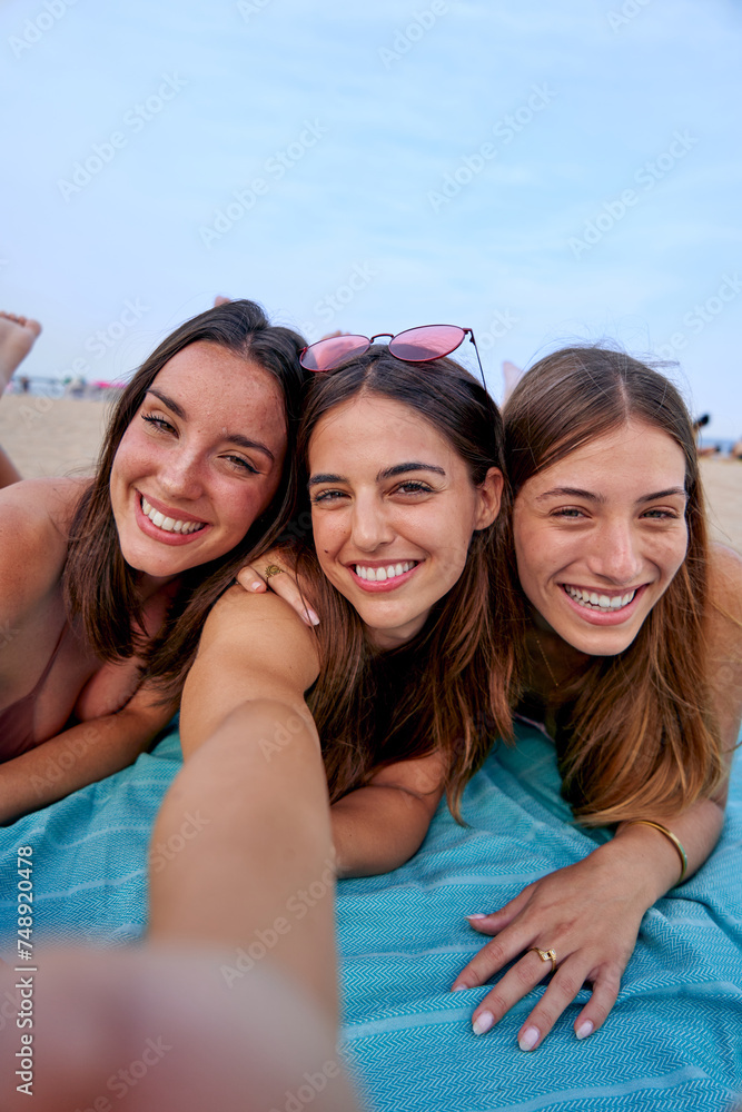 Three cheerful women lying on a towel using a cell phone to take vertical selfies for social media. Friends enjoying a beach vacation together. Three cheerful women lying on a towel using a cell phone to take vertical selfies for social media. Friends enjoying a beach vacation together.