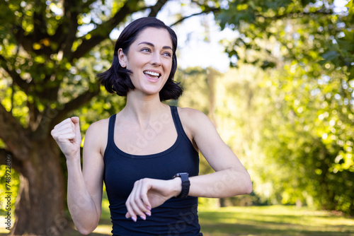 Fototapeta Naklejka Na Ścianę i Meble -  Close-up photo of happy young woman doing sports and jogging in park, looking at smart watch and showing success gesture with hand, rejoicing in victory