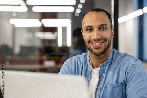 Close-up portrait of young smiling male freelancer and student working and studying in modern office, sitting at laptop and looking at camera photo