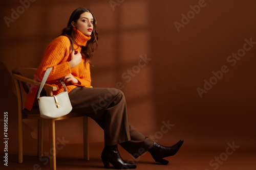 Fashionable confident woman wearing orange sweater, classic trousers, ankle boots, with white leather bag, posing on brown background. Studio fashion portrait. Copy, empty, blank space for text

