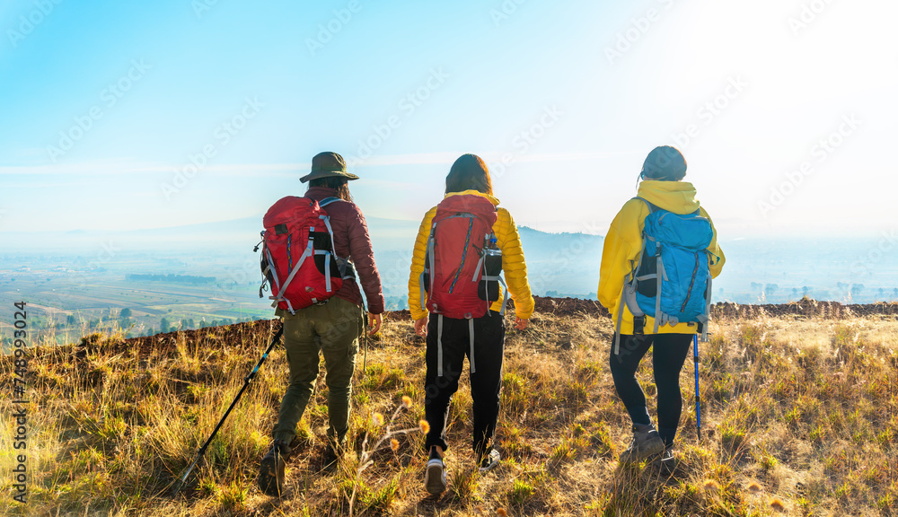 Three people walk up the mountain with hiking backpacks and yellow jackets, enjoying the sunrise and nature on their early hike.