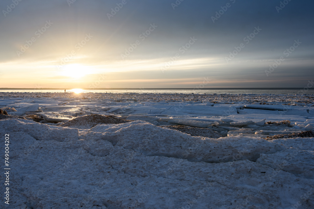 The Baltic Sea coast at the end of winter. The seashore covered with broken ice floes, in the evening golden sunlight. Bright sun in a cloudy hazy sky. Backlight