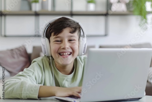 A young boy with a gray sweatshirt and white headphones uses a laptop, evidently amused by the content photo