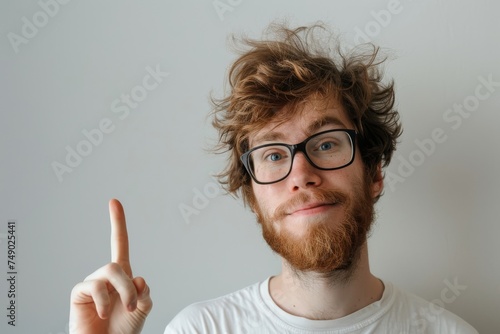 A young man with glasses and a beard pointing up  looking curious  against a plain backdrop