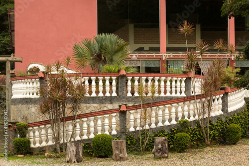 Casa com varanda, jardim com plantas e cerca viva e escada com guarda-corpo em concreto.  photo