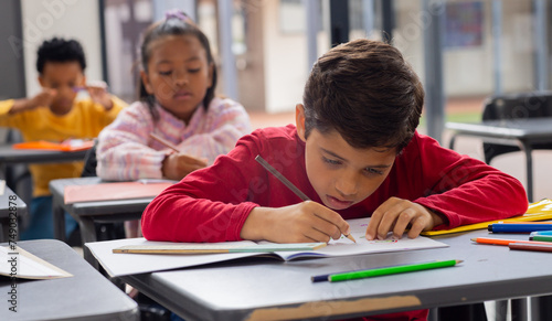 Biracial boy in red focuses intently on his drawing in a school classroom setting