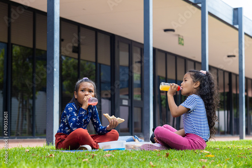 Two biracial girls enjoy lunch in school, sitting on the grass with sandwiches and juice