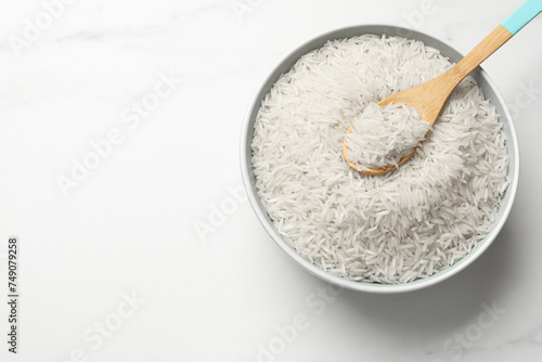 Raw basmati rice with spoon in bowl on white marble table, top view. Space for text photo