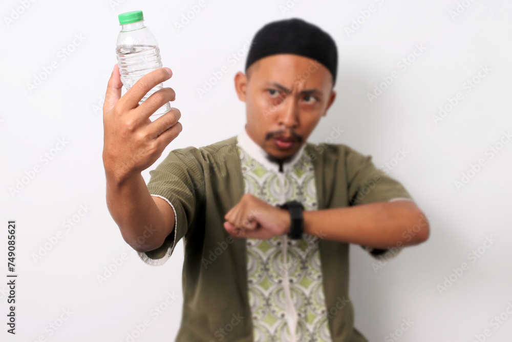 An Indonesian Muslim man in koko and peci checks his watch, waiting for iftar during the Ramadan fast. He holds a water bottle, indicating his thirst. Isolated on a white background An Indonesian Muslim man in koko and peci checks his watch, waiting for iftar during the Ramadan fast. He holds a water bottle, indicating his thirst. Isolated on a white background