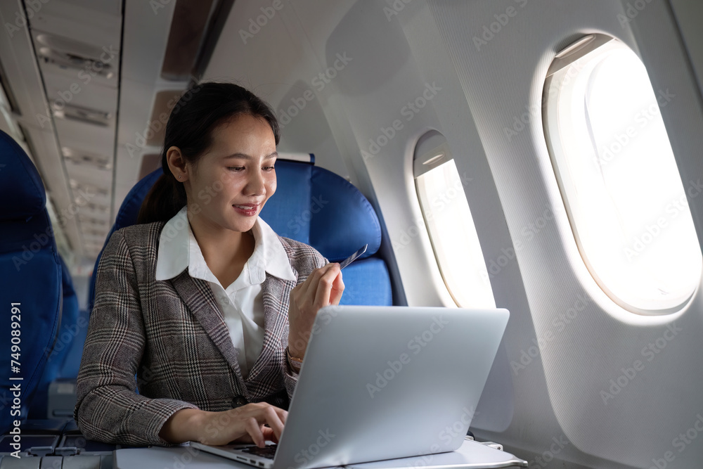 Attractive Asian female passenger on airplane sitting in comfortable seat using laptop and credit card, shopping using wireless connection, traveling in style
