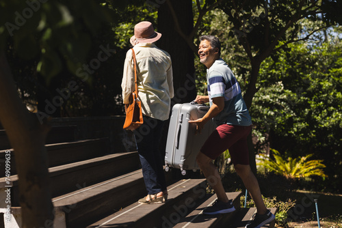 Senior biracial woman and biracial man ascend outdoor steps, man pulling a suitcase