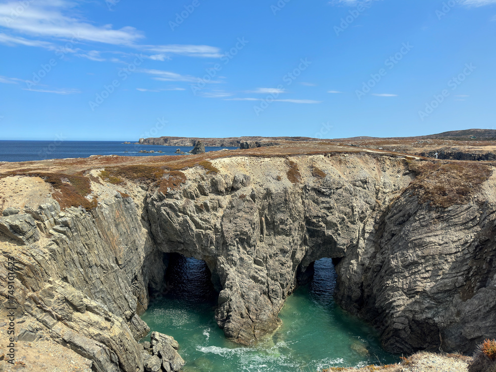A double rock arch formation with a large heart-shaped hole. The water is a green-teal color between the arches and the beach. Above the rocky cliff is covered in tundra-type ground covering plants. 