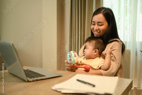 Smiling mom working with laptop and playing with her baby boy in living room