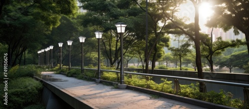 A metal bridge walkway in the middle of a park, with solar panels and garden lamps nearby. The walkway is illuminated by focused LED lights powered by sunlight. © AkuAku