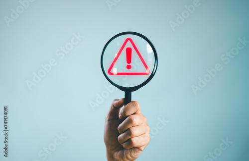 Hand holding magnifying glass with red triangle caution warning sign for notification error and maintenance concept.
