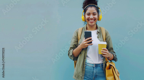 A cheerful student with headphones smiles while holding a smartphone and a coffee cup, standing against a blue background with a yellow backpack.