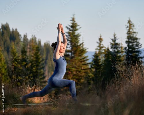 Sporty woman exercising outdoor. Young female doing warrior one pose. Workout on fresh air with view of mountains.