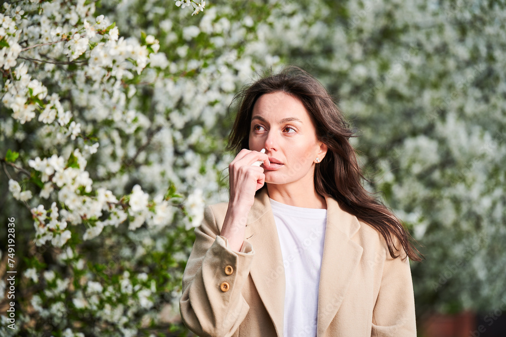 Woman allergic using medical nasal drops, suffering from seasonal allergy at spring in blossoming garden. Young woman treating runny nose in front of blooming tree outdoors. Spring allergy concept. Woman allergic using medical nasal drops, suffering from seasonal allergy at spring in blossoming garden. Young woman treating runny nose in front of blooming tree outdoors. Spring allergy concept.