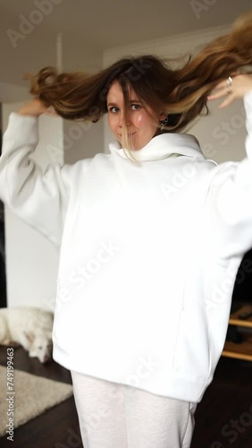 beautiful girl in a white hooded sweatshirt dresses and tries on clothes. vertical video. a girl near the TV and a white shepherd dog.