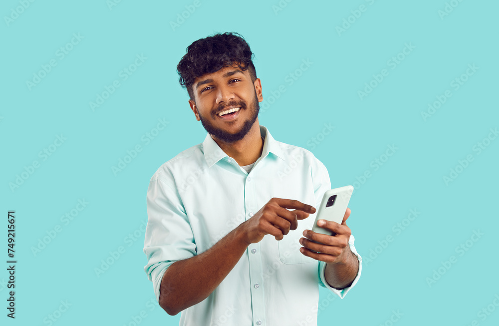 Young happy Indian man holding mobile phone and smiling looking at camera using application for managing business or company dressed in white shirt stands in studio on turquoise background Young happy Indian man holding mobile phone and smiling looking at camera using application for managing business or company dressed in white shirt stands in studio on turquoise background