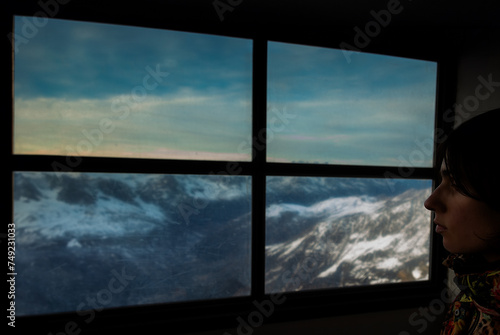 A young woman gazes wistfully out a rustic ski chalet window at dusk, her silhouette framed against the fading light on snow-capped French mountains.