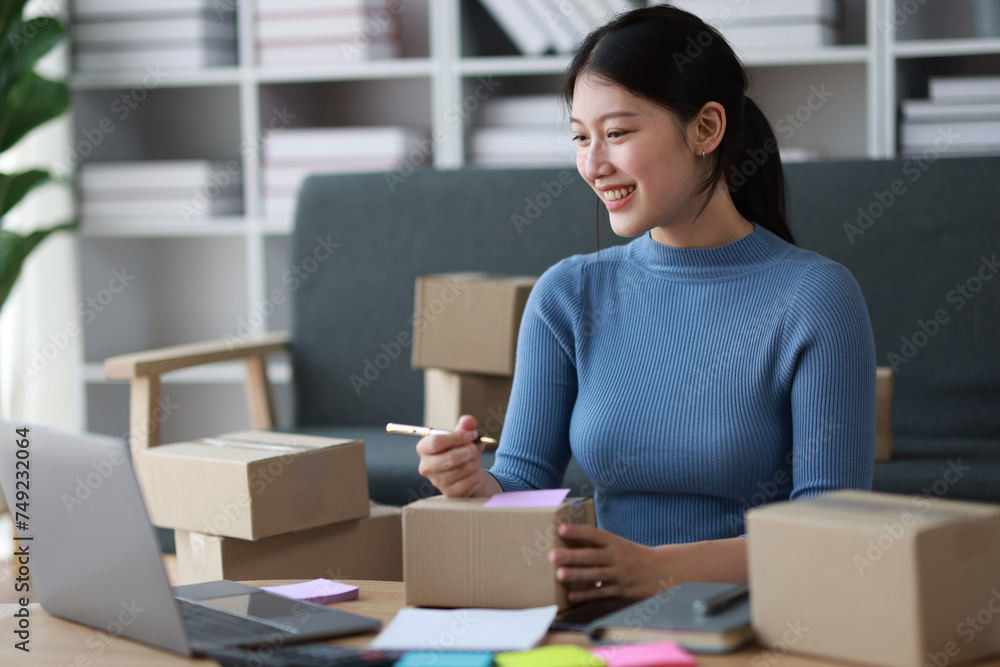 Young Asian woman working with parcel box at home preparing to deliver goods and writing online order note from internet on laptop. Young Asian woman working with parcel box at home preparing to deliver goods and writing online order note from internet on laptop.