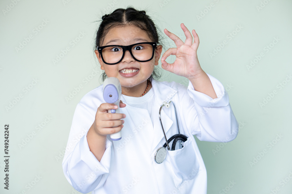Cute girl in a medical mask plays a doctor and measures the temperature with a non-contact thermometer and showing like ok signs. Cute girl in a medical mask plays a doctor and measures the temperature with a non-contact thermometer and showing like ok signs.