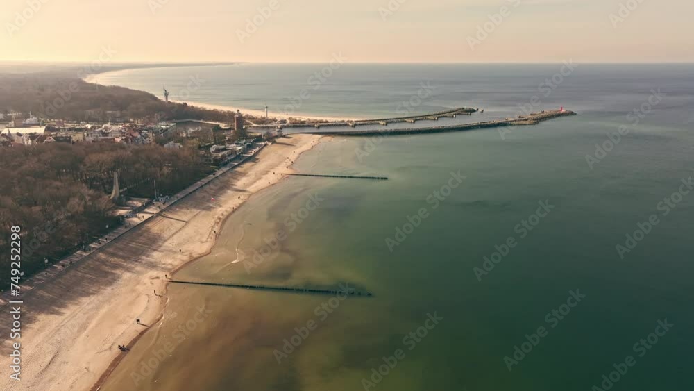 Drone captures sunny Kołobrzeg in February, with tourists strolling, wide sandy beach, calm Baltic Sea, seagulls, lighthouse, and port entrance. Drone captures sunny Kołobrzeg in February, with tourists strolling, wide sandy beach, calm Baltic Sea, seagulls, lighthouse, and port entrance.