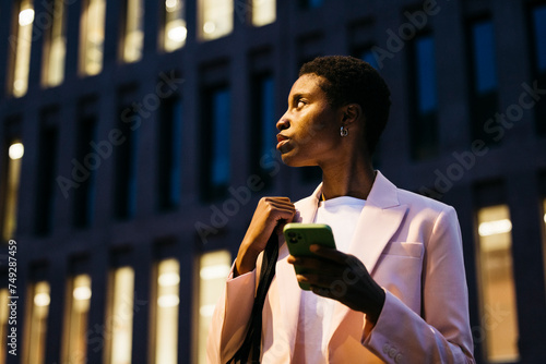 Young black stylish businesswoman with a phone on her hand looking away. Businesswoman with a mobile phone on downtown disctrict at night. photo