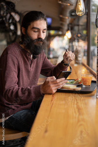 Hipster bearded man eating his lunch sitting in cafe and use mobile phone for texting sms, reading news