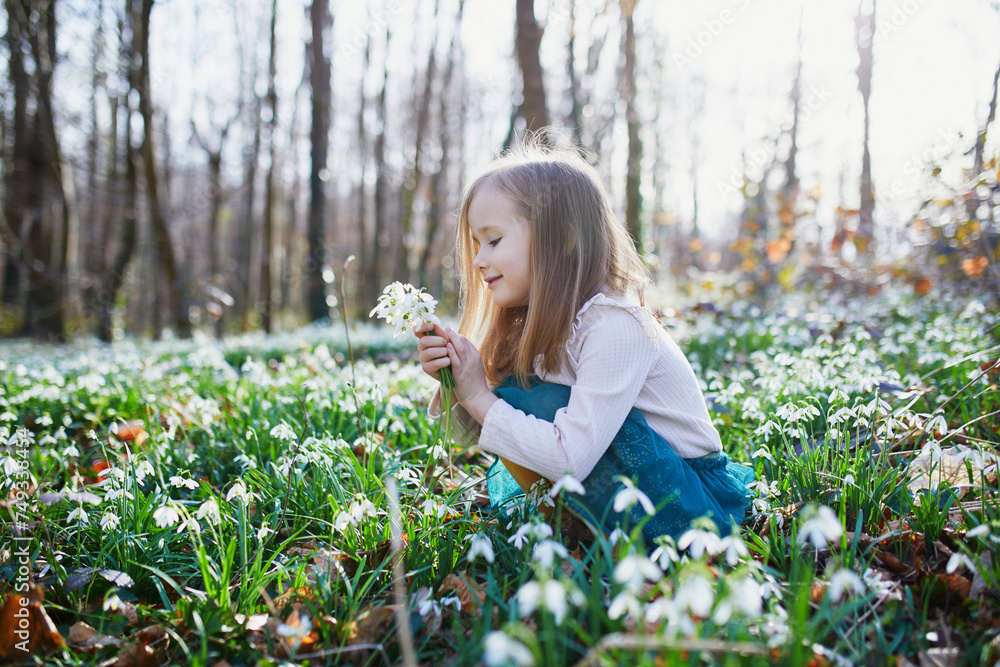 Cute preschooler girl in green tutu skirt gathering snowdrop flowers in park or forest on a spring day