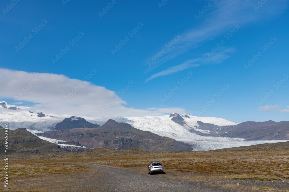 Panoramic view of the Fjallsarlon glacier in Iceland seen from a dirt road close to the terminus of the glacier with a car parked on the road against a blue sky with some feather clouds Panoramic view of the Fjallsarlon glacier in Iceland seen from a dirt road close to the terminus of the glacier with a car parked on the road against a blue sky with some feather clouds