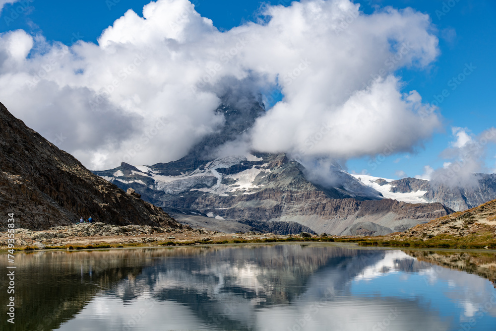 Panoramic view over the water of the Riffelsee near Zermatt, Switzerland towards the Matterhorn partly covered in white clouds, also reflected in the water of the lake; part of the Monte Rosa massif