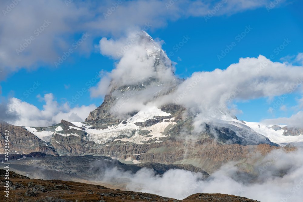 Low angle view of near symmetric pyramidal peak of the Matterhorn near Zermatt, Switzerland in extended Monte Rosa area of the Pennine Alps somewhat surrounded by white clouds in otherwise blue sky Low angle view of near symmetric pyramidal peak of the Matterhorn near Zermatt, Switzerland in extended Monte Rosa area of the Pennine Alps somewhat surrounded by white clouds in otherwise blue sky