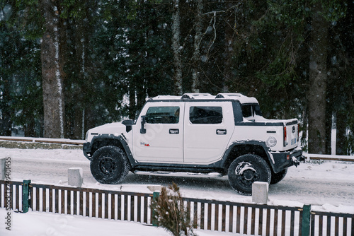 White Hummer H2 pickup truck drives under snowfall along a snow-covered road along a coniferous forest. Side view