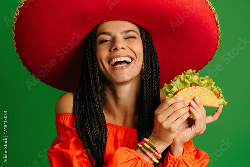 Happy young Mexican woman in Sombrero holding taco and smiling against green background